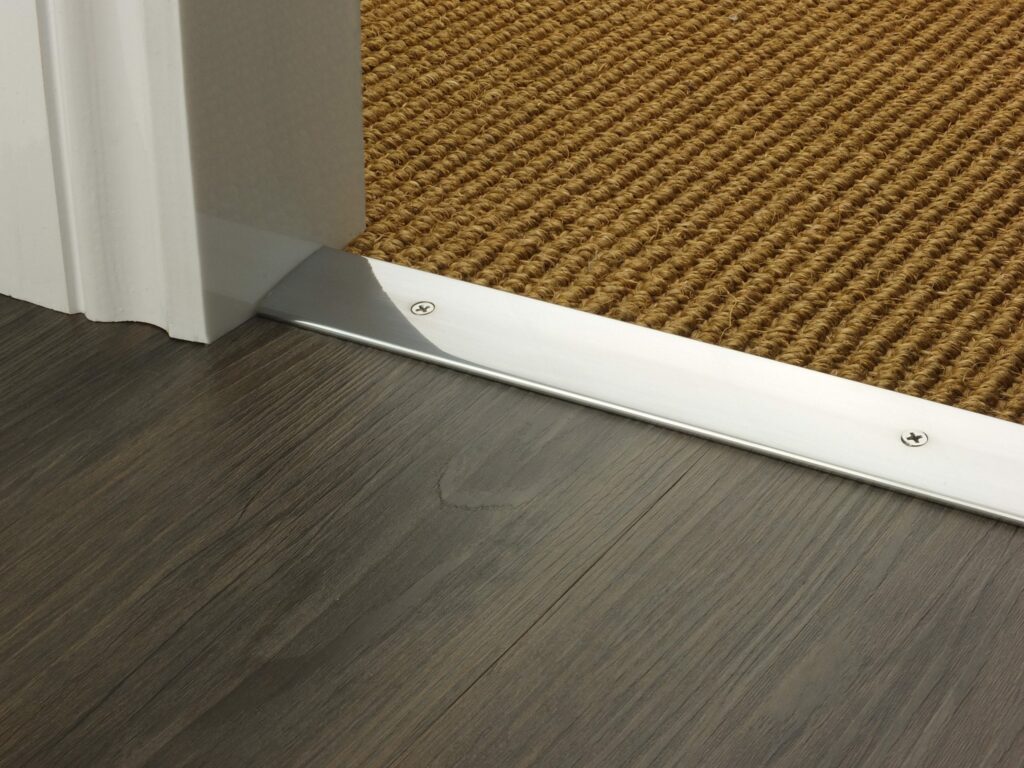 Threshold plate in polished chrome with visible screws joins cord carpet to brown vinyl floor